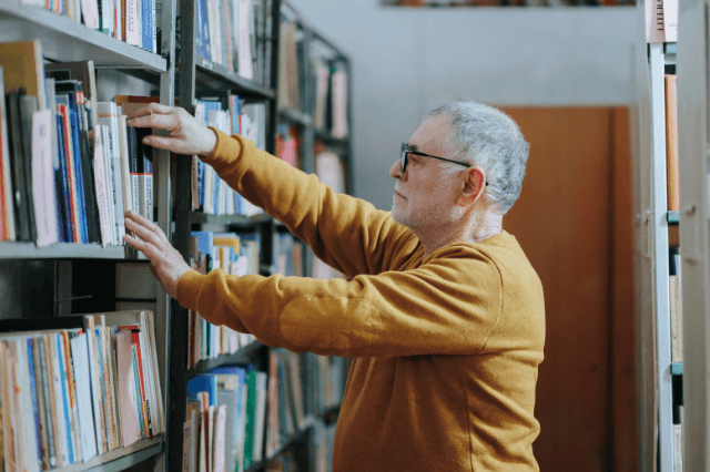 A man in his early 60s looking at a bookshelf with a calm, analytical expression, illustrating a healthy cognitive state during normal aging.