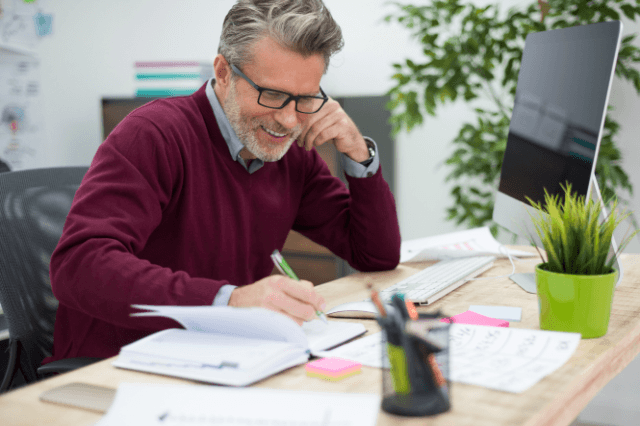 A candid, warm-lit photograph of a distinguished man in his late 60s, wearing glasses, deeply focused on studying from a book and taking notes at a modern desk. A laptop is open beside him. He has an expressions of engagement and mental clarity, illustrating what a sharp mind feels like and cognitive longevity in daily life. Professional home office environment.